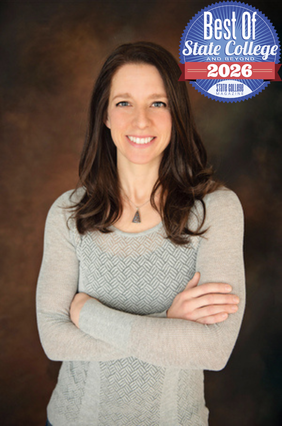 A woman with long brown hair smiling, wearing a light gray patterned sweater and a necklace, posed with arms crossed against a dark, soft-focus background.