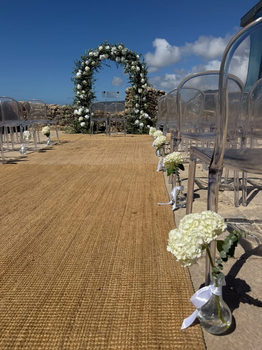 A wedding ceremony setup with a natural fiber aisle runner, clear chairs on each side decorated with white flowers, and a floral arch backdrop under a blue sky with some clouds.