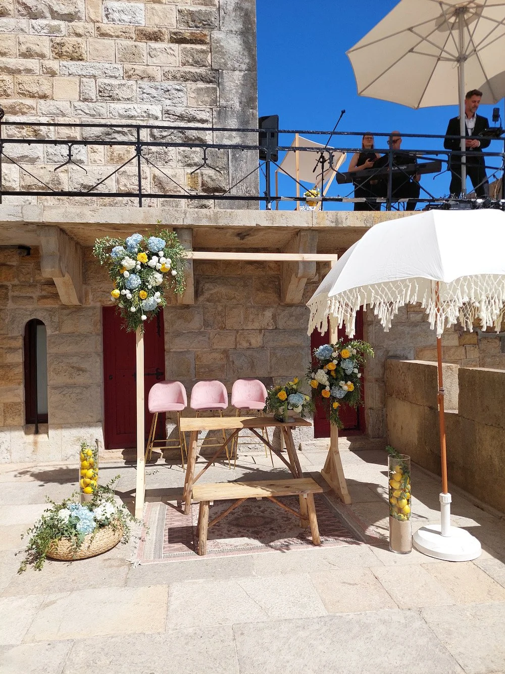 Wedding setup with pink chairs, floral arrangements, a white umbrella, and a wooden table against a stone building with a balcony and live music band.