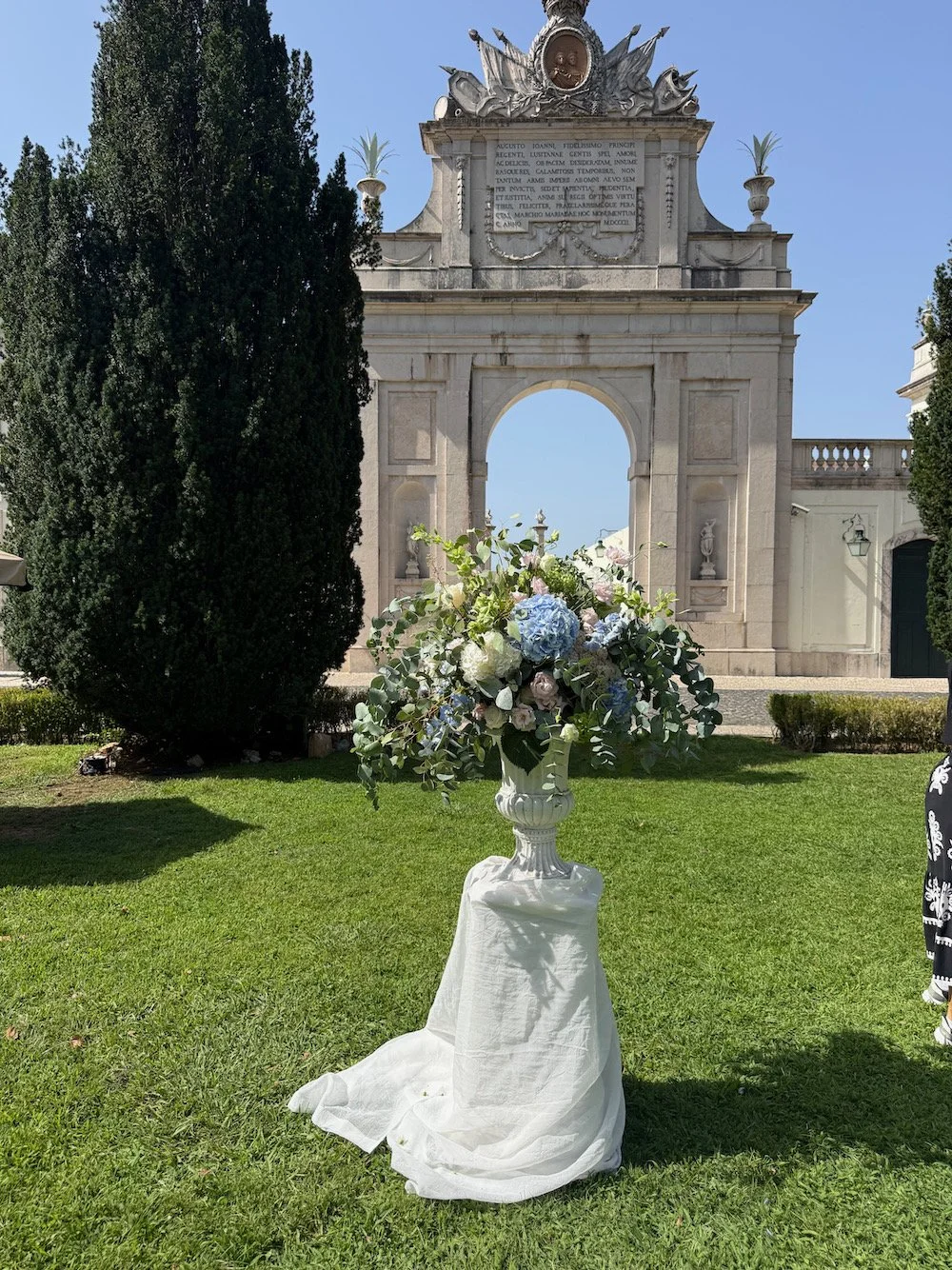 A floral arrangement in a white vase with blue and white flowers on a table covered with white cloth, in front of a historic stone archway decorated with statues and Latin inscriptions, under a clear blue sky.