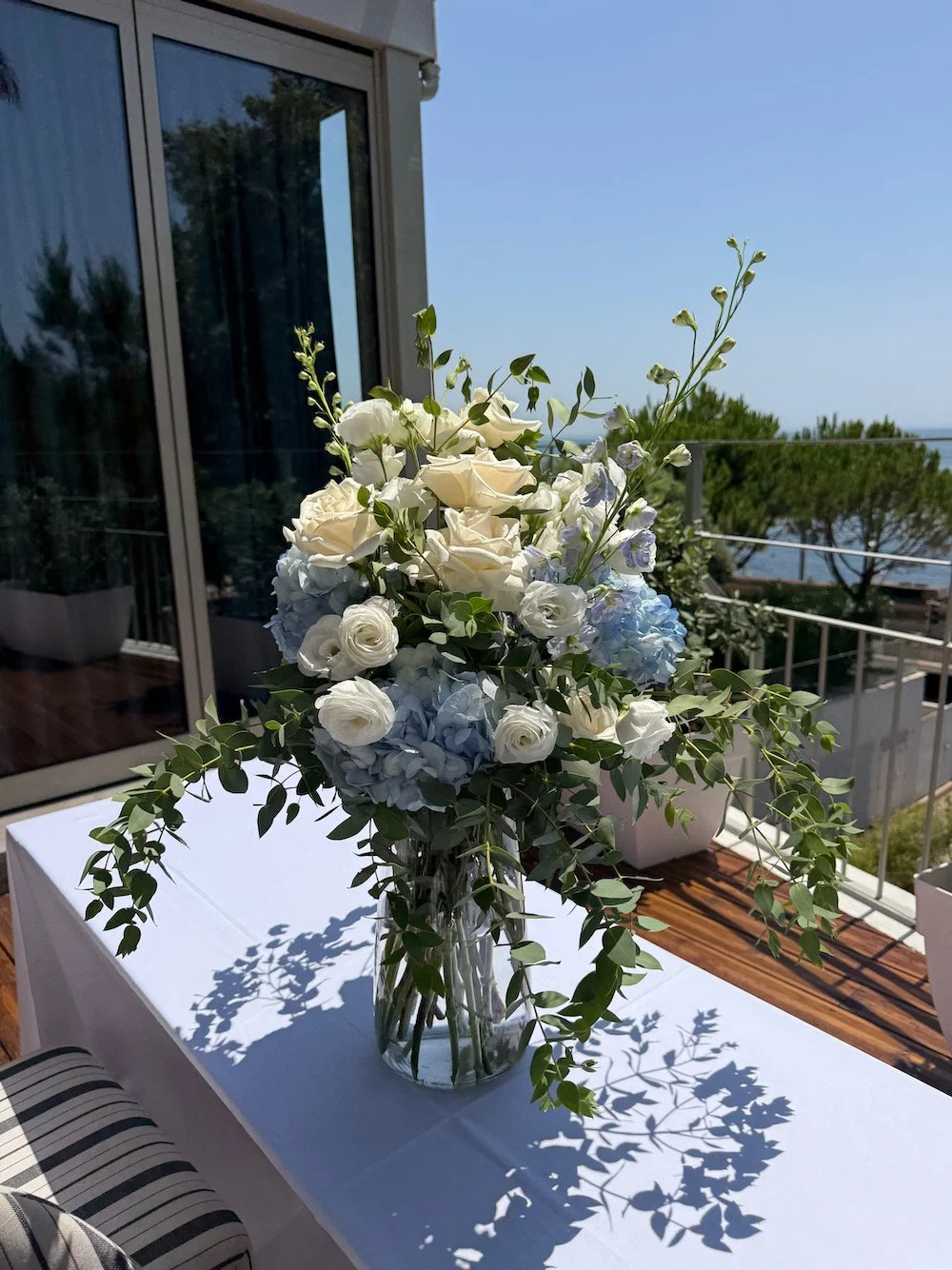 A tall glass vase filled with white roses, light blue hydrangeas, and greenery, placed on a white table on a balcony with a view of trees, water, and a clear blue sky.