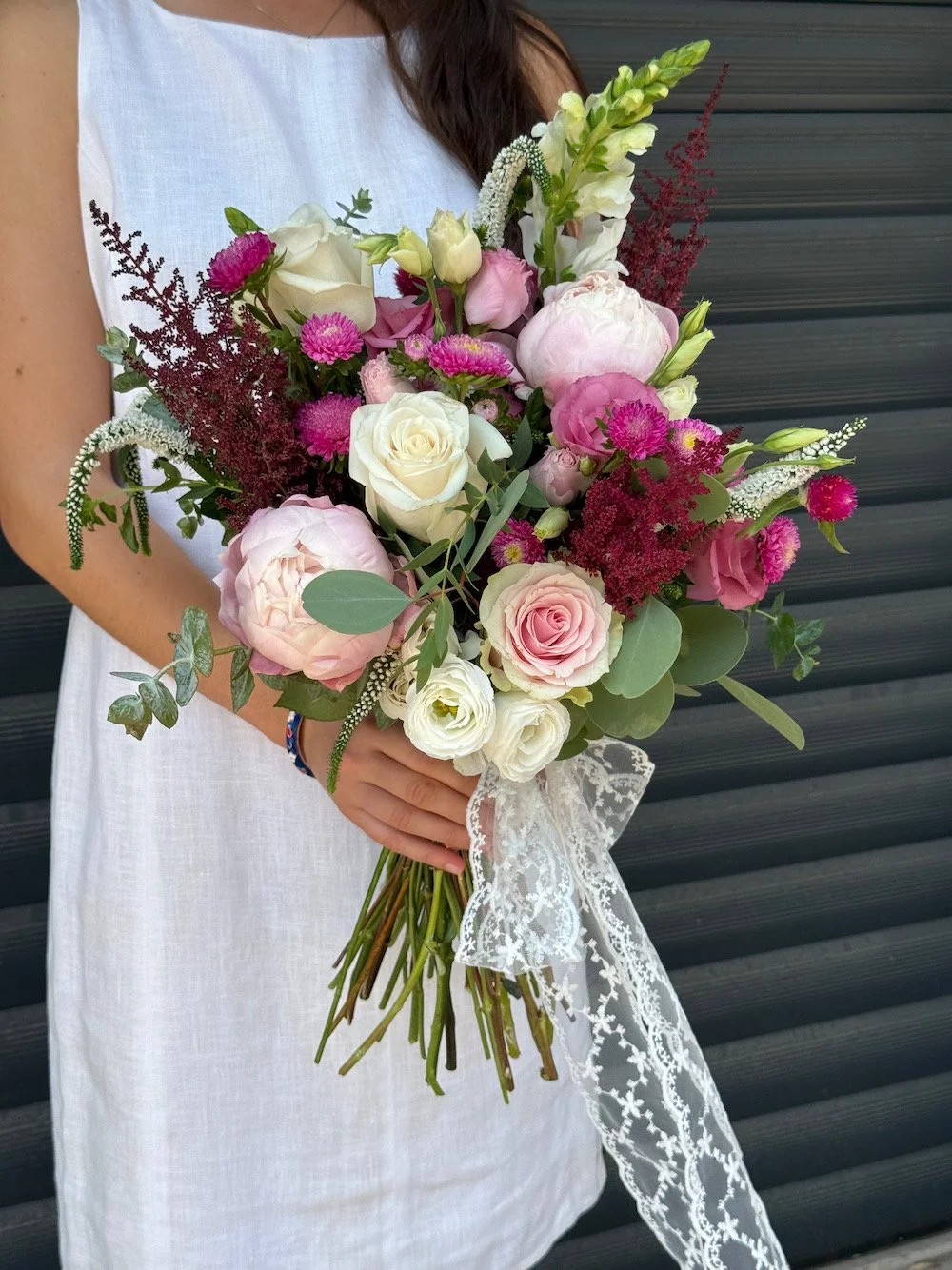 Person holding a bouquet of pink, white, and cream flowers with greenery, tied with a lace ribbon, standing in front of a dark, horizontal siding background.