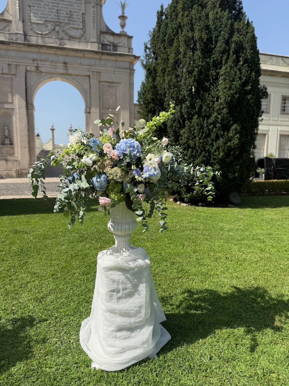 A large floral arrangement in a white urn on a pedestal, featuring white, pink, and blue flowers with greenery, set outdoors on a manicured lawn with a historical stone building and trees in the background.