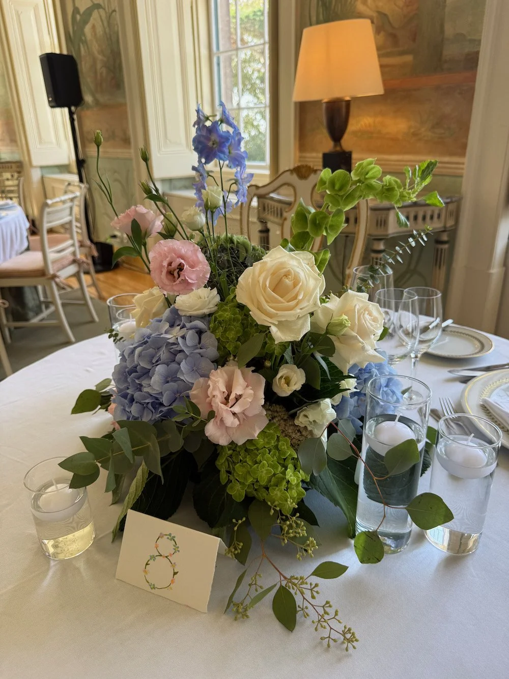 A floral centerpiece with white roses, pink and blue flowers, green foliage, on a round table with place settings including wine glasses, candles, and a table number card in an elegant dining room.