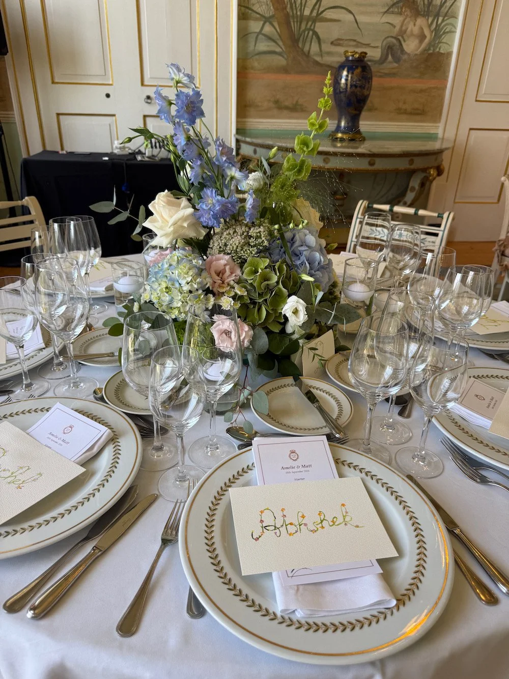 Elegant round dining table set with white and gold china, silverware, multiple wine glasses, and a floral centerpiece with blue, white, and pink flowers. Place cards and napkins are arranged at each setting.