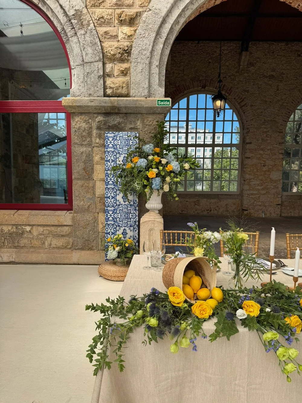 Elegant indoor event space decorated with floral arrangements, including a table with yellow roses, white flowers, and greenery, and large vases of flowers against exposed brick and stone walls.