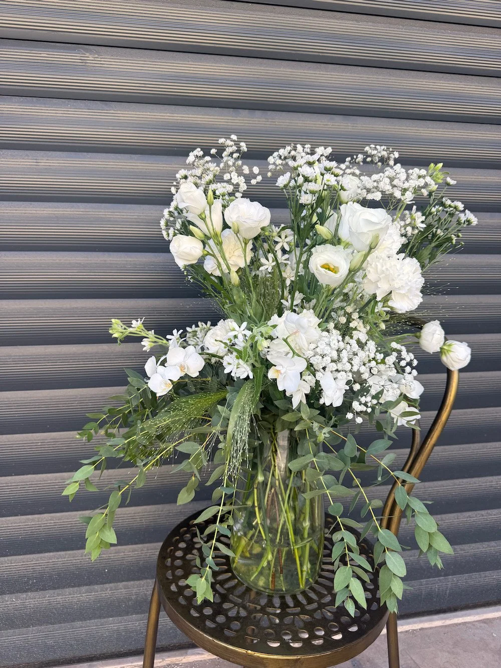 A bouquet of white flowers in a glass vase on a black metal chair with a decorative seat, background of gray horizontal metal siding.