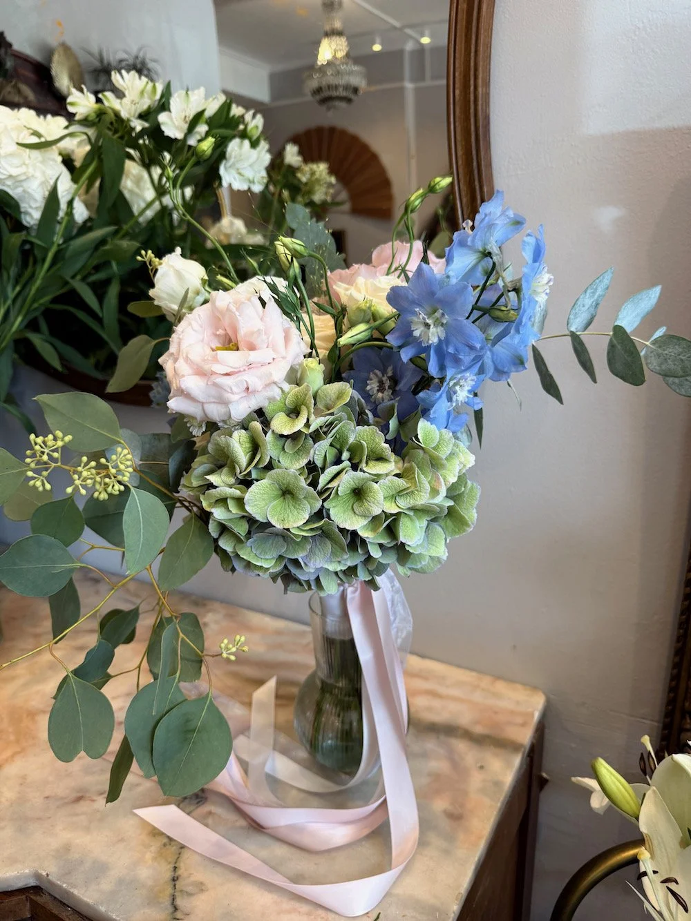A bouquet of light pink, white, and blue flowers with green leaves in a glass vase on a marble table.
