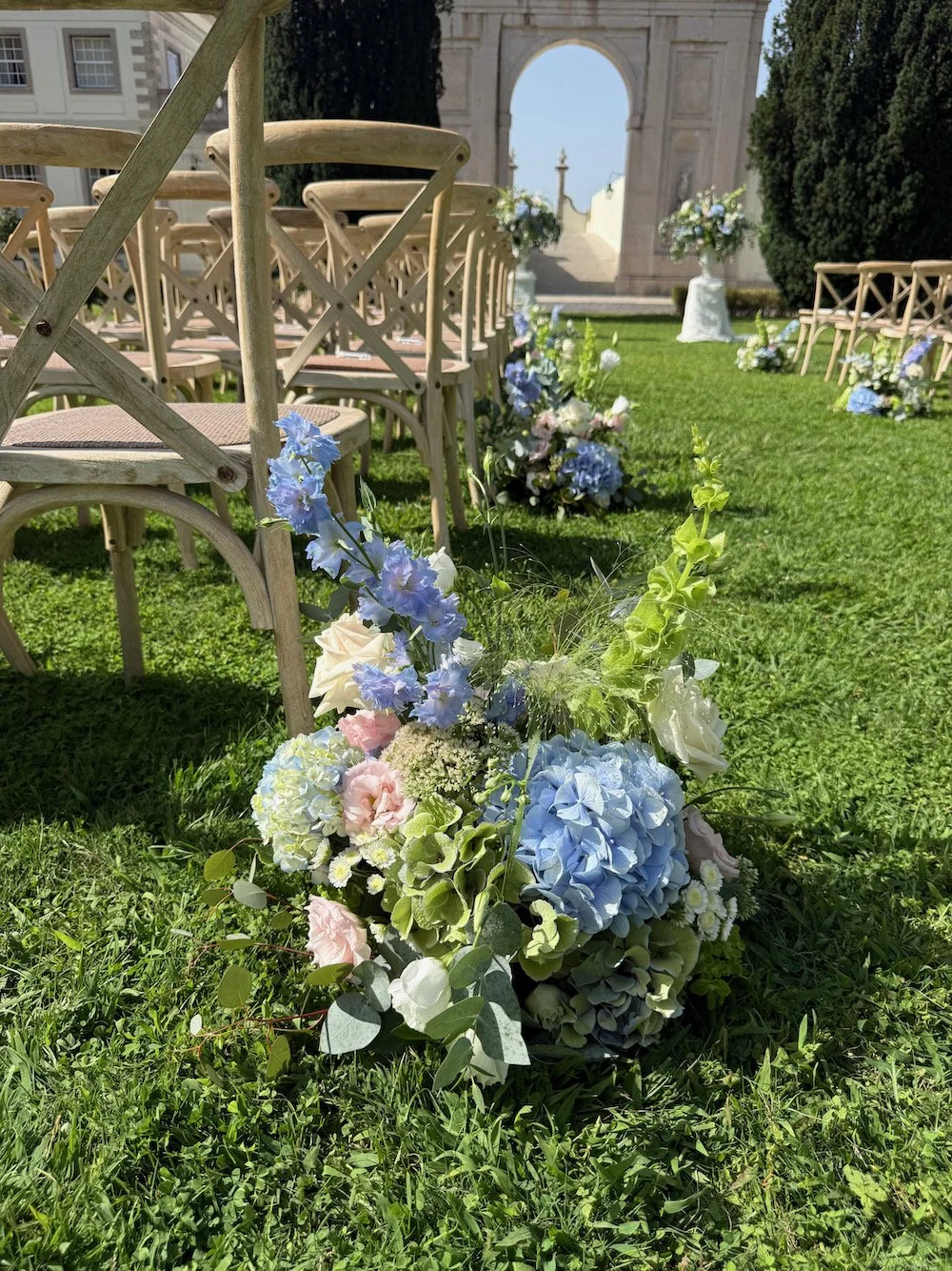 Outdoor wedding setup with rows of wooden chairs, floral arrangements, and an archway in the background.