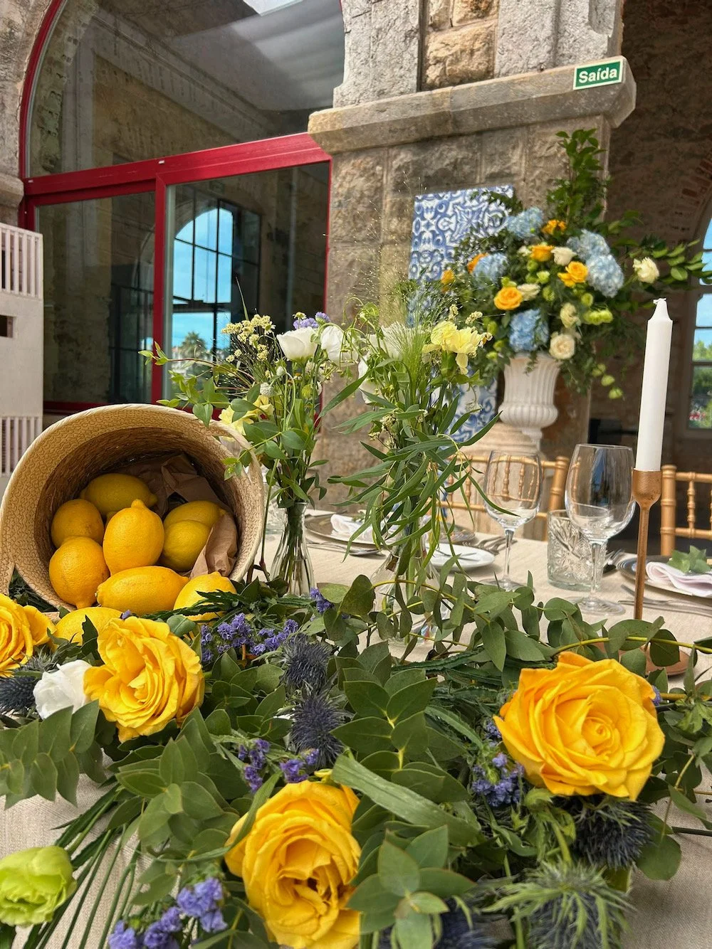 A table decorated with vibrant yellow roses, purple flowers, and green foliage, with a basket of lemons, set in a rustic stone-walled room with large windows and a floral arrangement in a white vase.