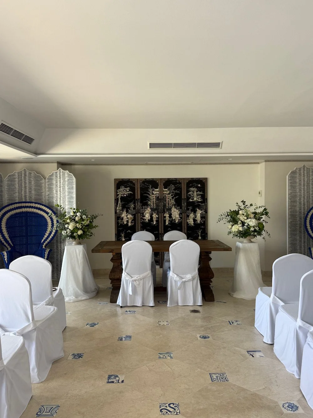 A decorated room with white chair covers, floral arrangements, and a dark wooden table, likely set up for a formal event or ceremony.