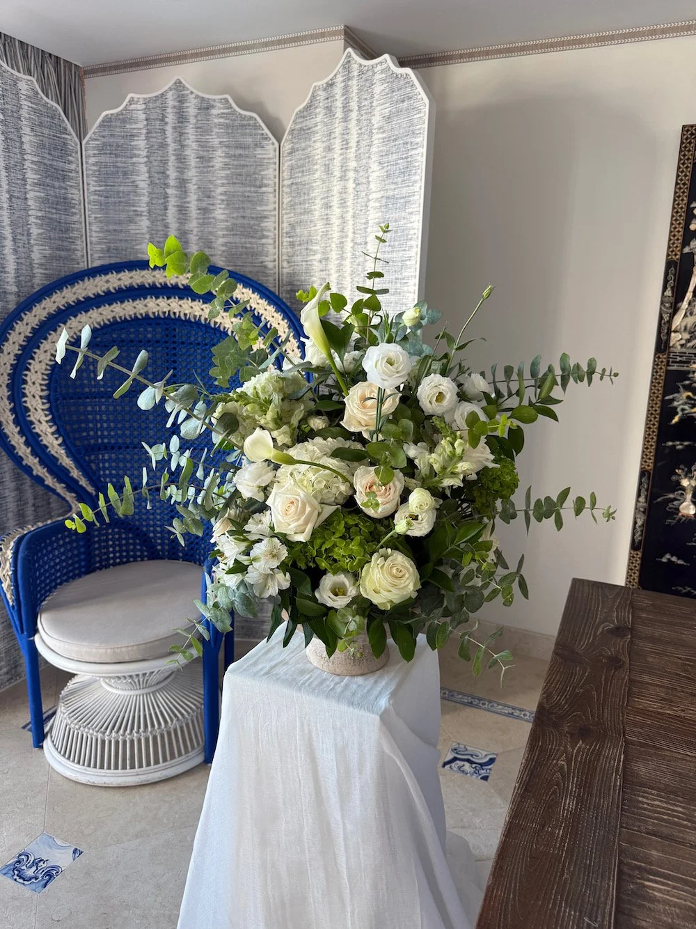 A large floral arrangement with white roses, lisianthus, and green foliage on a white tablecloth in a room with a blue wicker chair and a decorative folding screen in the background.
