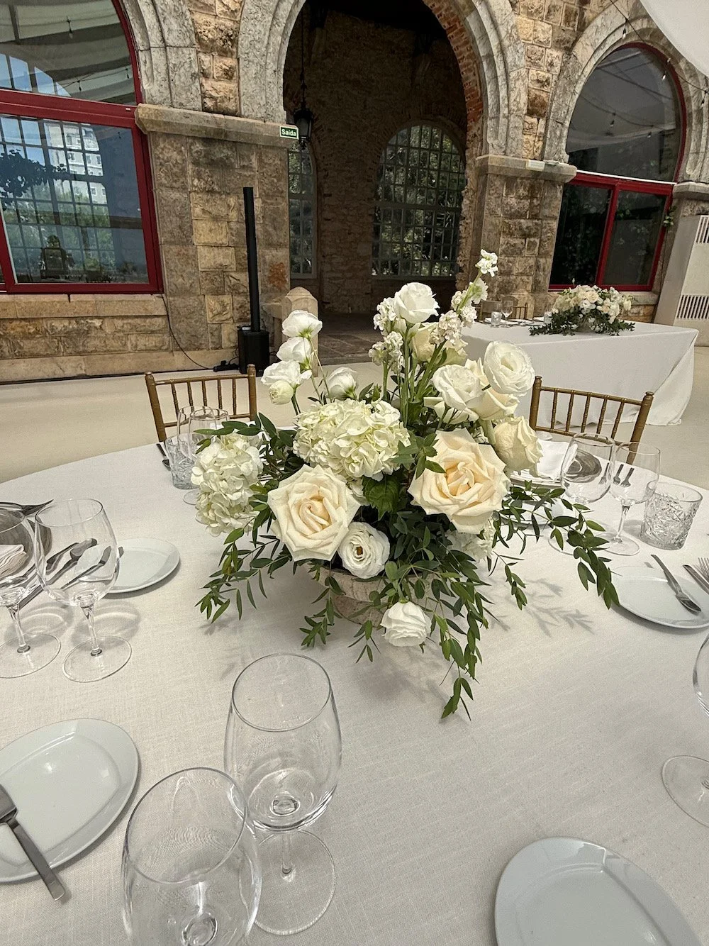 Elegant round table set for a formal event, decorated with a white tablecloth, a large floral centerpiece with white roses and greenery, surrounded by wine glasses, water glasses, plates, and cutlery in a rustic venue with stone walls and large windo