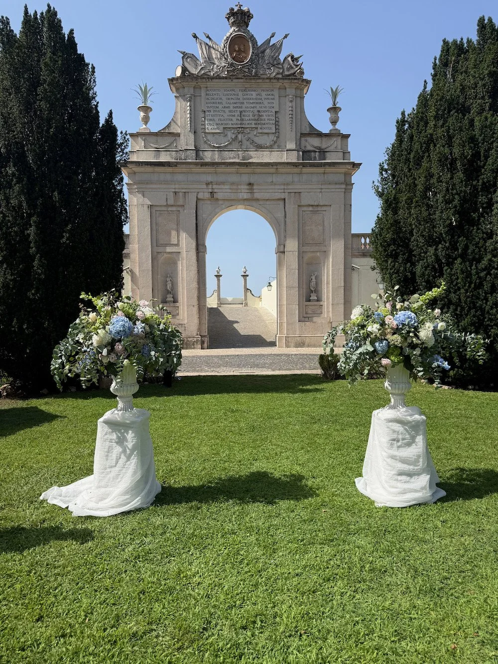 Wedding site with two floral arrangements in white urns on a green lawn, stone archway with Latin inscription in the background, tall trees on both sides, and a clear blue sky.