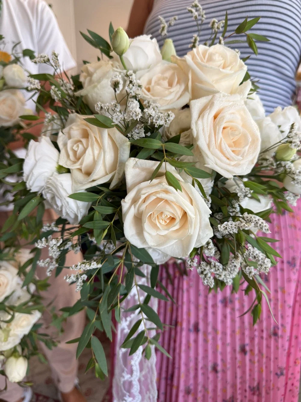 A bouquet of white roses with greenery and small white flowers, held by a person wearing a striped shirt.