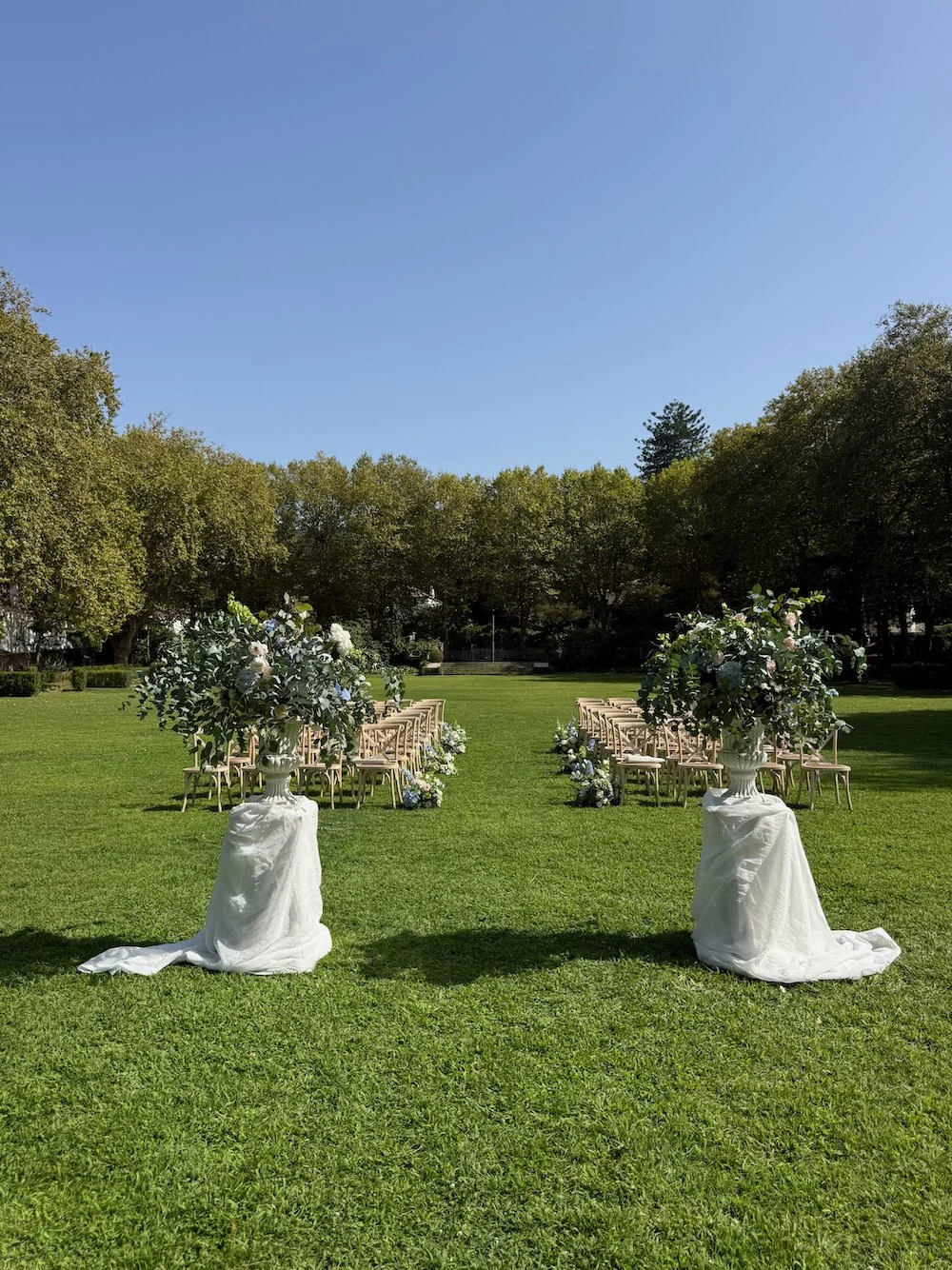 Outdoor wedding ceremony setup on a grassy field with two flower arrangements in white urns at the entrance, rows of chairs decorated with flowers, and trees in the background under a blue sky.