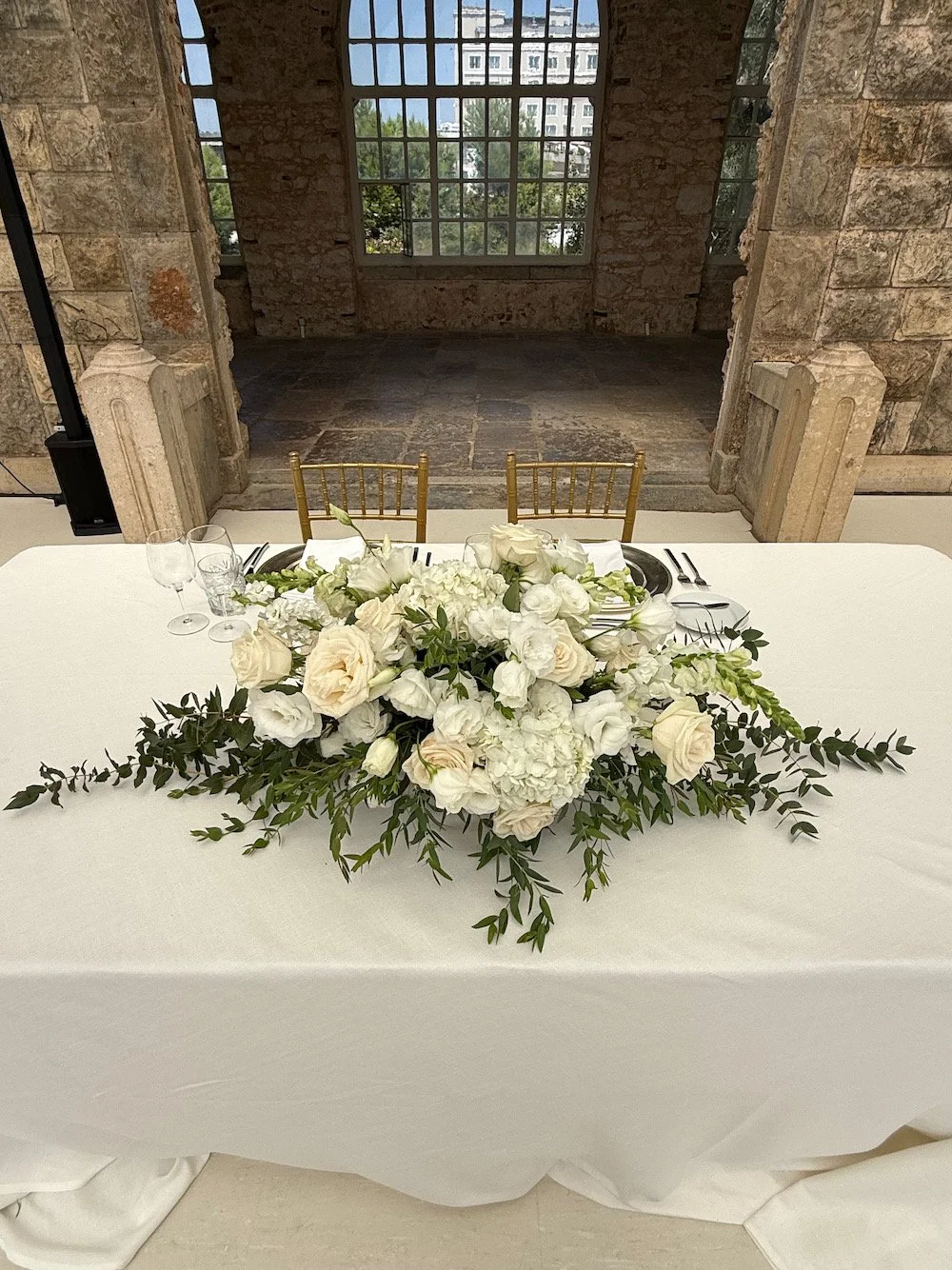 A white tiered table set for a formal event with a large white floral centerpiece with roses and greenery, surrounded by gold chairs, against rustic brick walls and large windows.