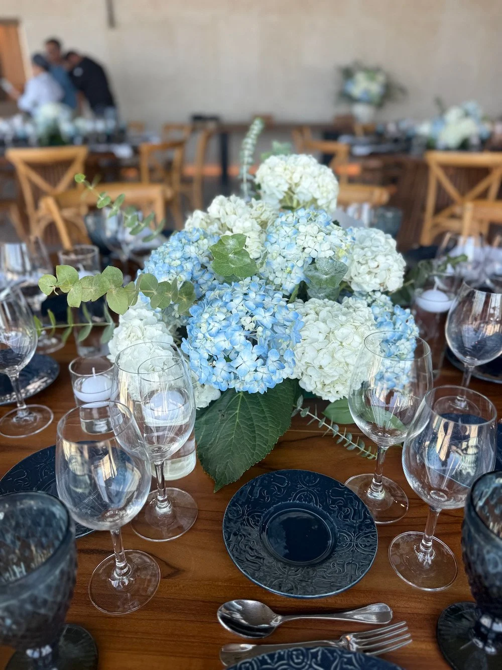 A table decorated with a floral centerpiece of white and blue hydrangeas, surrounded by empty glasses, dark plates, and cutlery, set for a formal event or celebration.