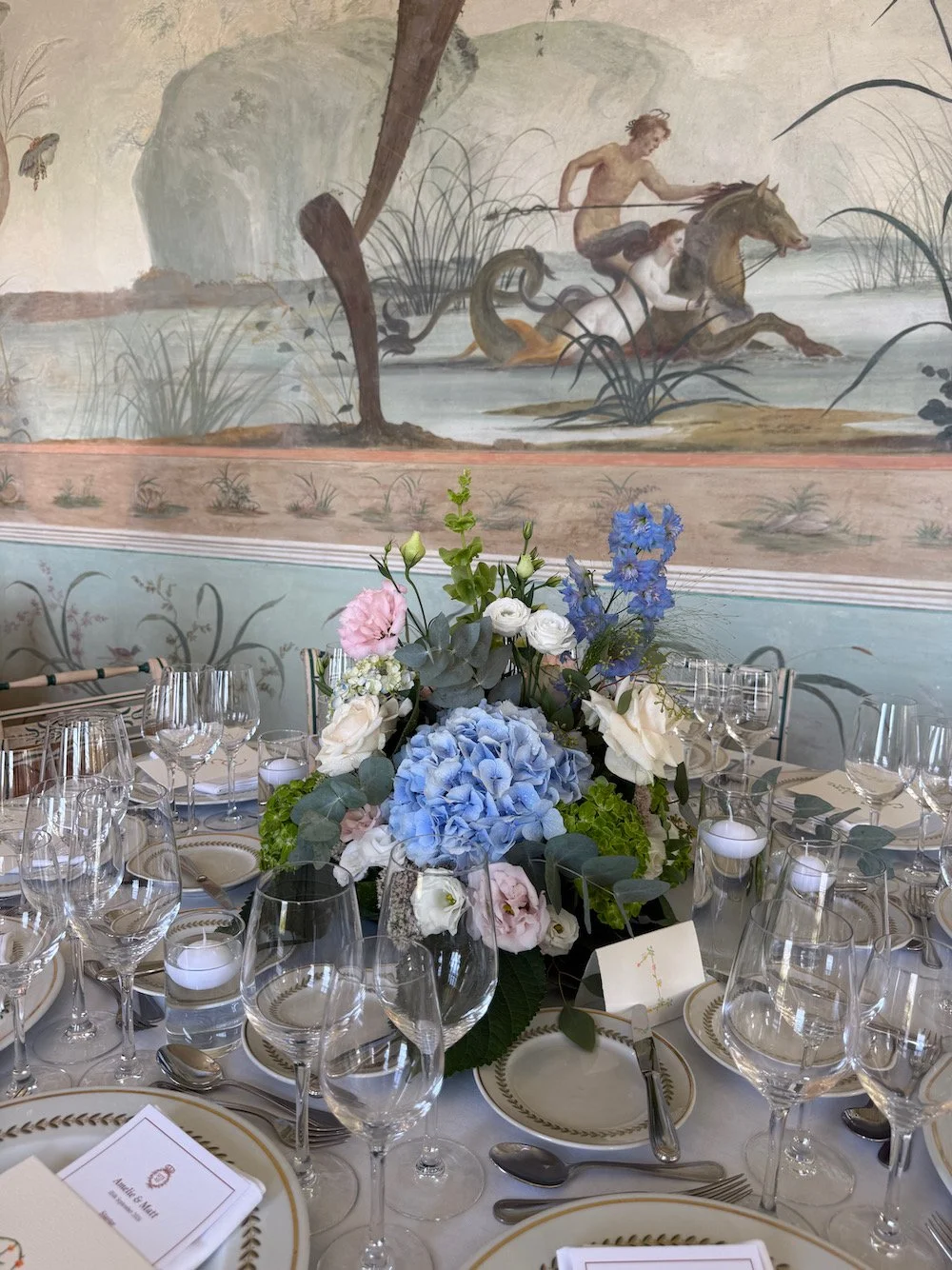 A banquet table set with glassware, plates, and silverware, featuring a floral centerpiece with blue and white flowers, in front of a mural depicting two mythological figures riding a horse and a serpent in a wetland scene.