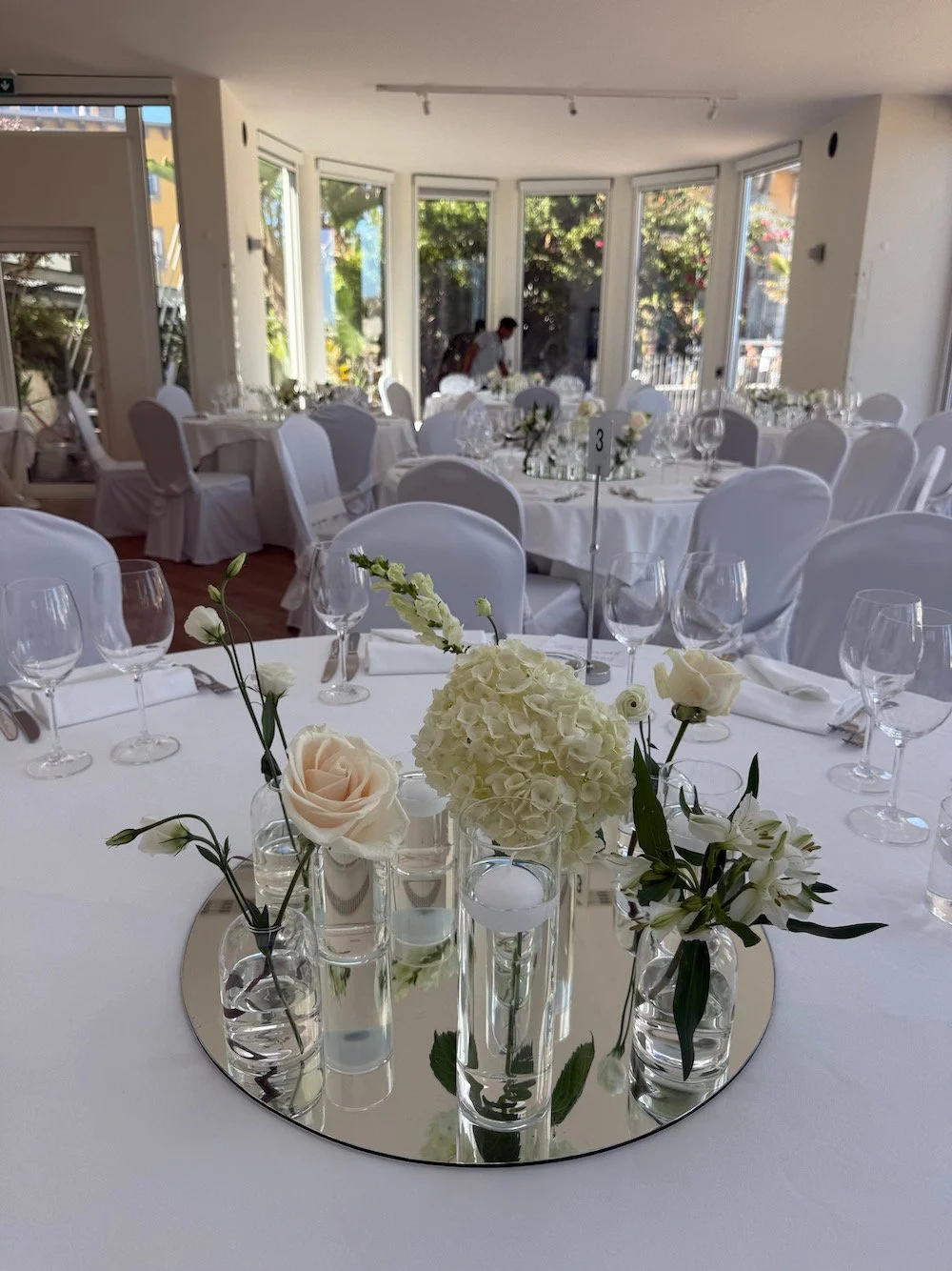 Elegant indoor wedding reception with white tablecloths, floral centerpieces, and glassware, set in a bright room with large windows showing greenery outside.