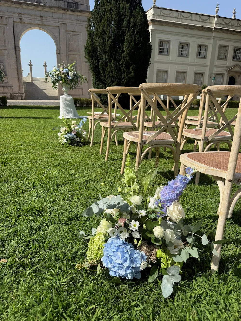 Outdoor wedding setup with wooden chairs arranged on a grassy lawn, decorated with blue, white, and green florals, facing an ornate stone archway and a classical building in the background.