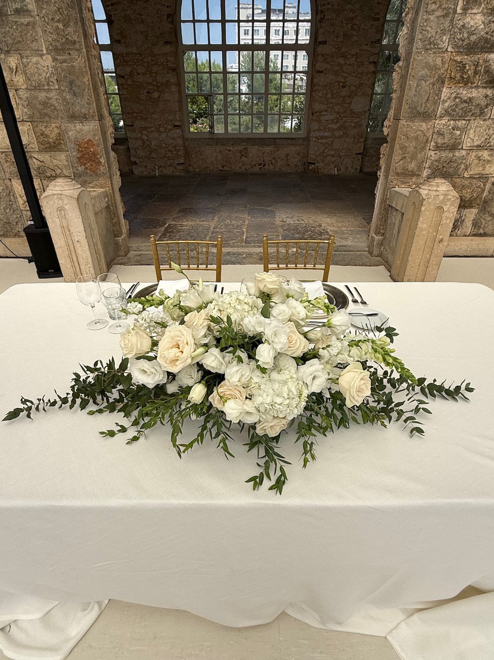 A wedding or event table with a large white floral centerpiece, set for guests with wine glasses, plates, and cutlery, in a historic building with stone walls and large windows.
