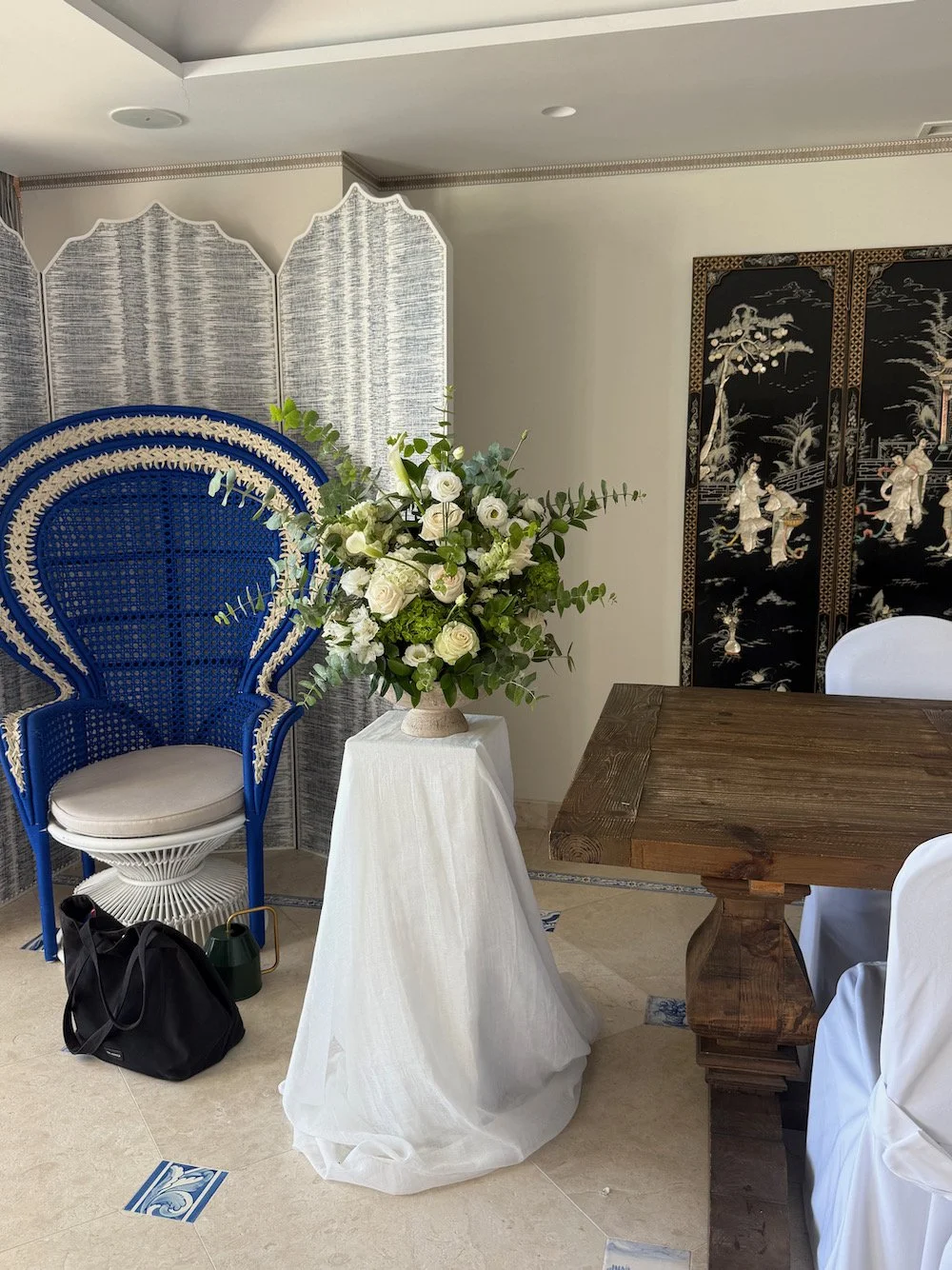 Interior of a room with a rustic wooden table, a large floral arrangement on a white draped stand, a blue and white decorative chair, and traditional Asian-style black folding screen with painted figures.