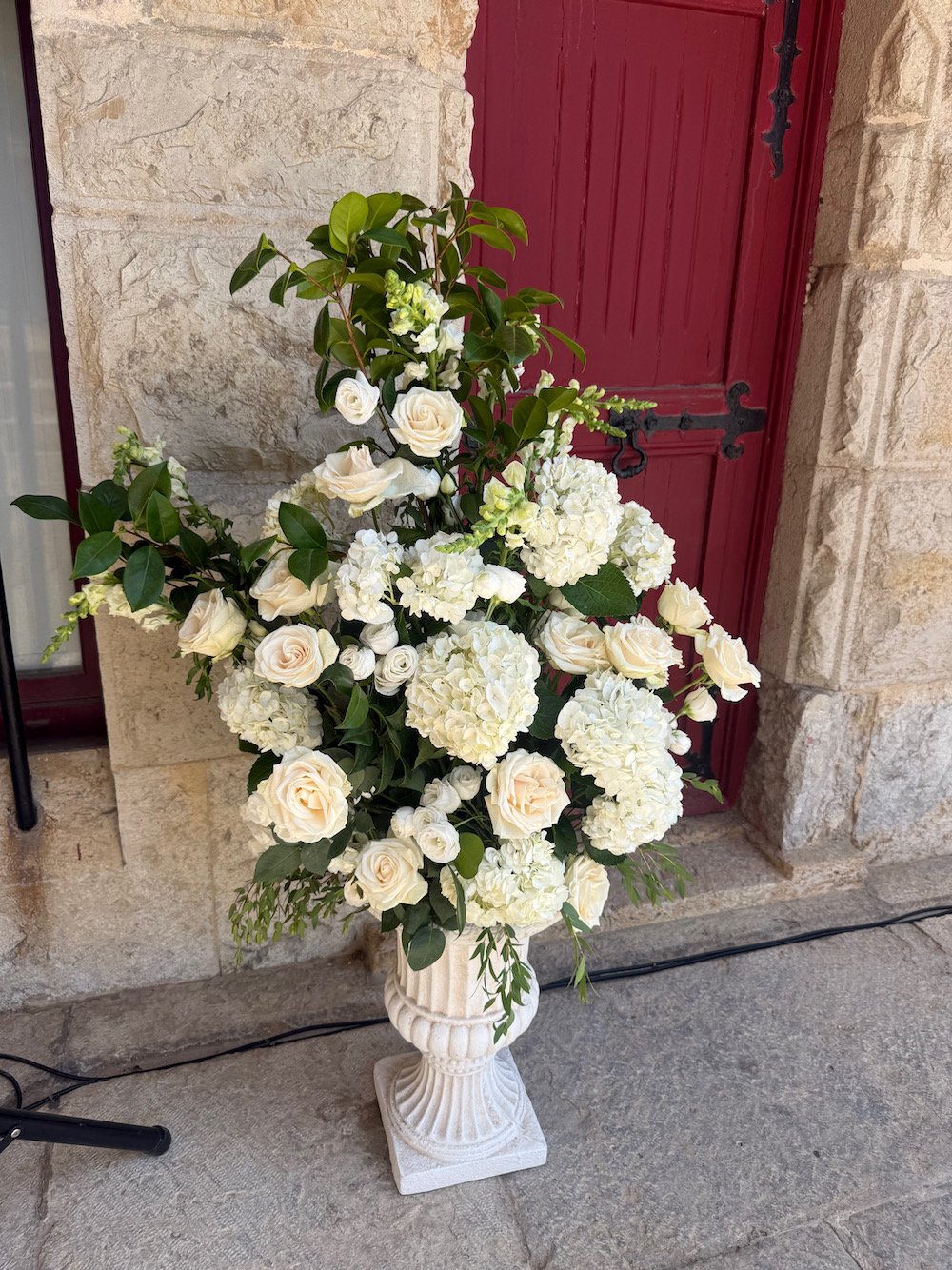 White floral arrangement with roses and hydrangeas in a decorative cream-colored pedestal vase, placed outdoors near a stone wall and red wooden door.