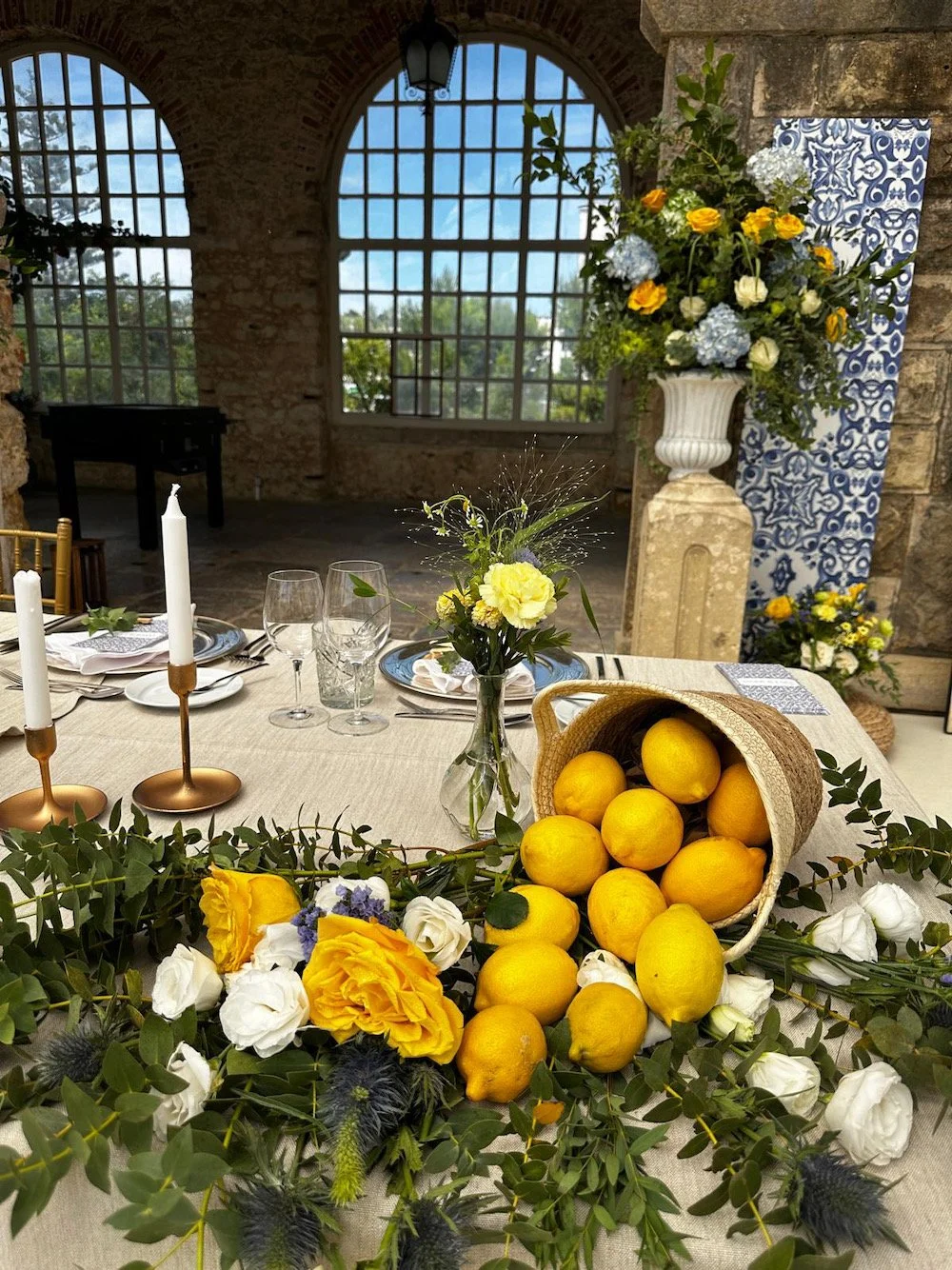 A table decorated for a formal event or celebration, with a centerpiece of assorted flowers, a basket of lemons, and elegant tableware. The background features large arched windows and a rustic brick interior.