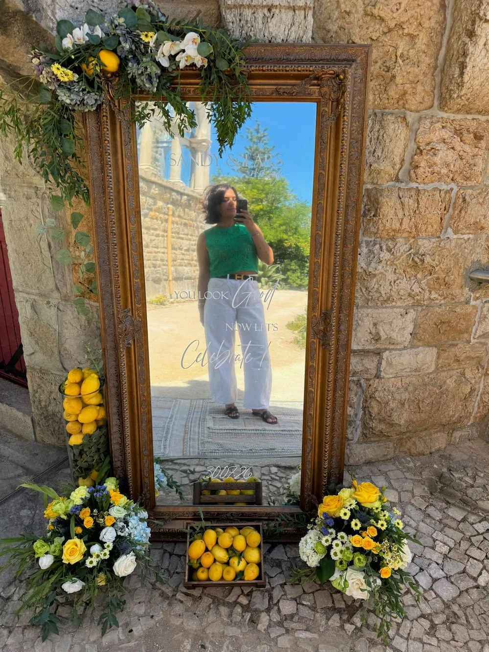 Person taking a selfie in front of a decorative outdoor mirror decorated with yellow lemons and floral arrangements, with a stone building and greenery in the background.