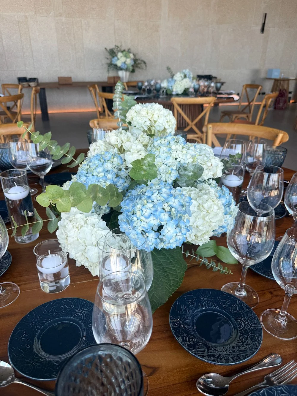 Elegant table setting with a floral centerpiece of white and light blue hydrangeas, surrounded by wine glasses, candles, and dark blue plates in a decorated event hall.