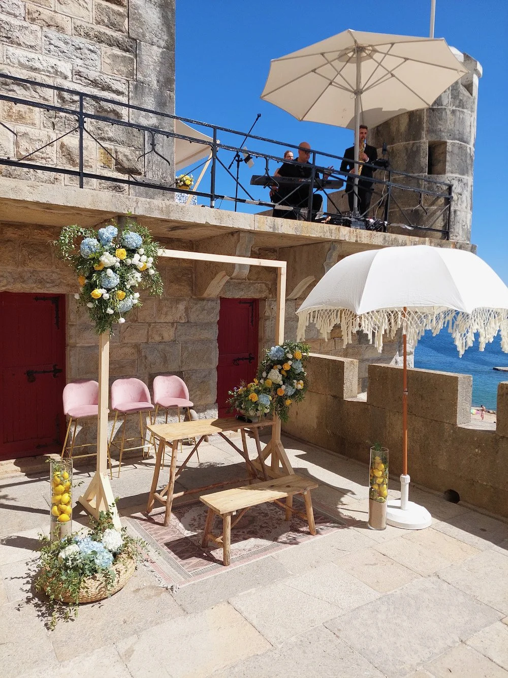 Decorated outdoor wedding setup with pink chairs, floral arrangements, wooden table and benches, and large umbrellas, on a stone terrace with castle-like stone walls overlooking the sea. Musicians are performing on a balcony above.