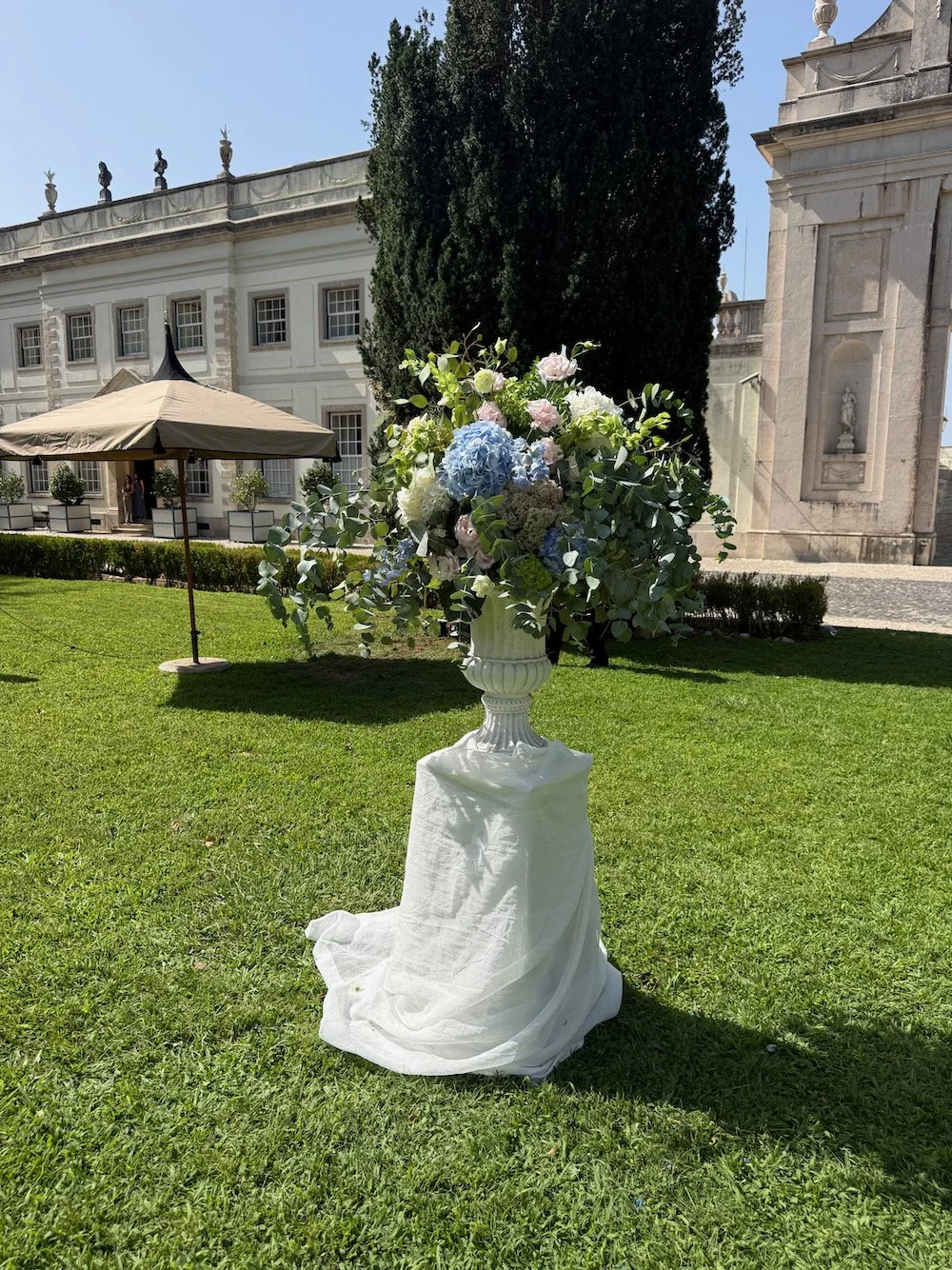 A large floral arrangement in a white pedestal vase sits on a draped white cloth on the grass. The arrangement includes blue, white, and pink flowers with green foliage. In the background, there is a historic white building, a large tree, a beige umb