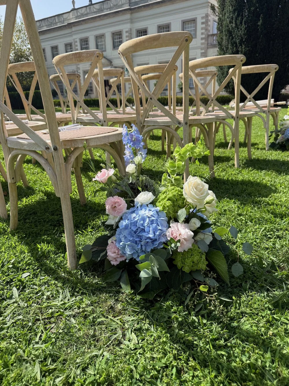 Empty outdoor chairs arranged on green grass in a garden or outdoor setting, with a bouquet of colorful flowers including blue hydrangeas, pink roses, and white flowers in the foreground, and a white building with large windows in the background.