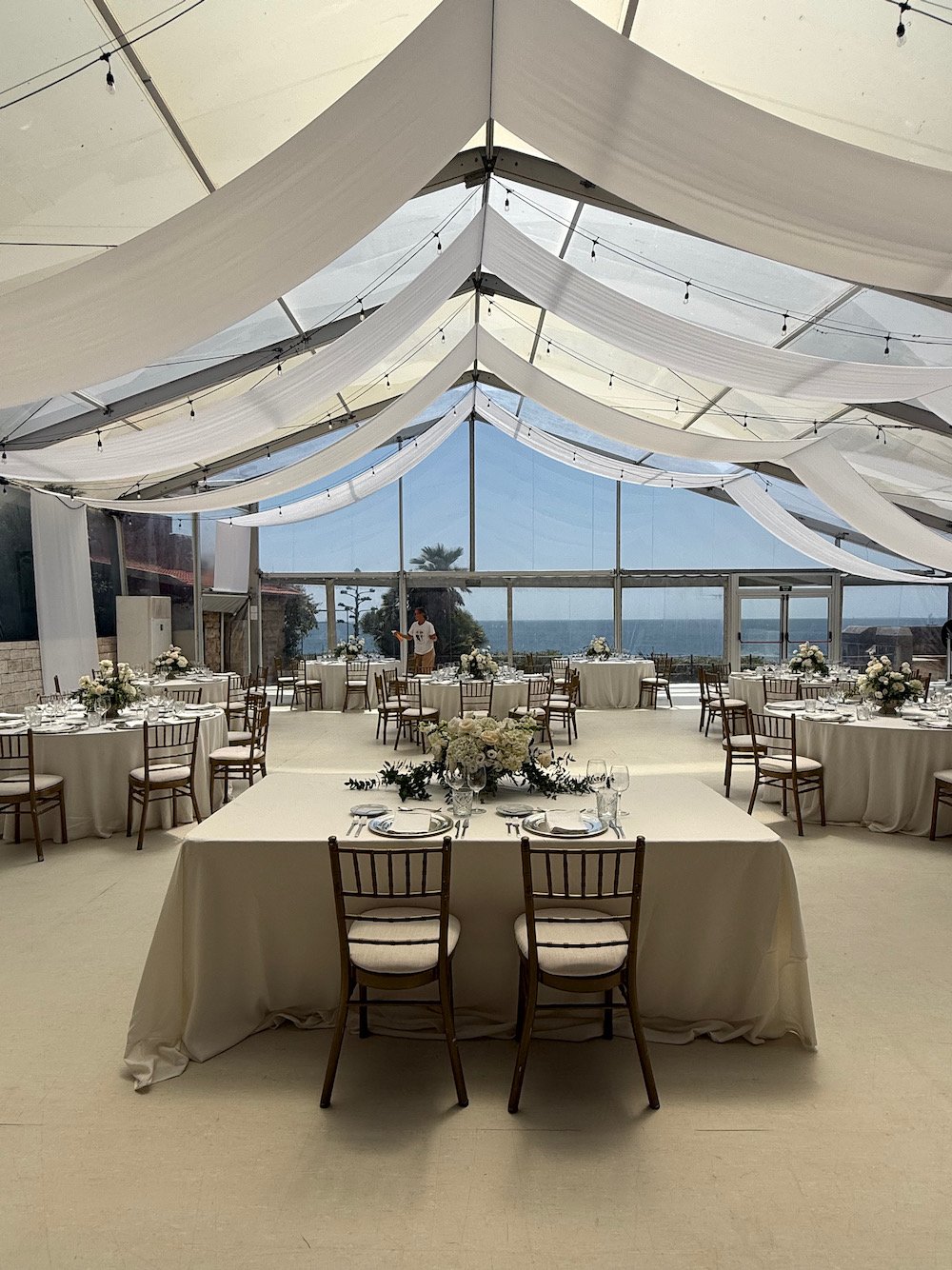Elegant indoor event space decorated with white draped fabric on the ceiling, large round tables with white tablecloths, floral centerpieces, and place settings, with floor-to-ceiling windows showing a view of the ocean and palm trees outside.