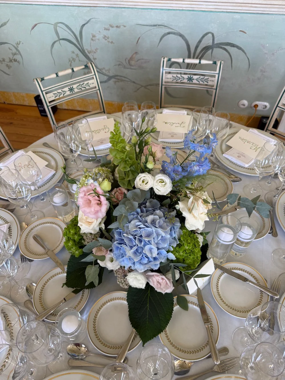 A round table set for a formal dinner with a centerpiece of blue, white, and pink flowers, surrounded by white plates with gold trim, and empty wine glasses.