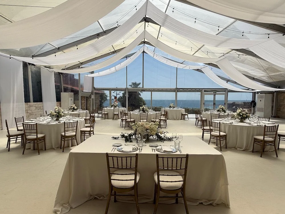 Elegant indoor event space with round tables, floral centerpieces, and a large window showing a view of the ocean and palm trees outside. Ceiling drapes and string lights decorate the space.