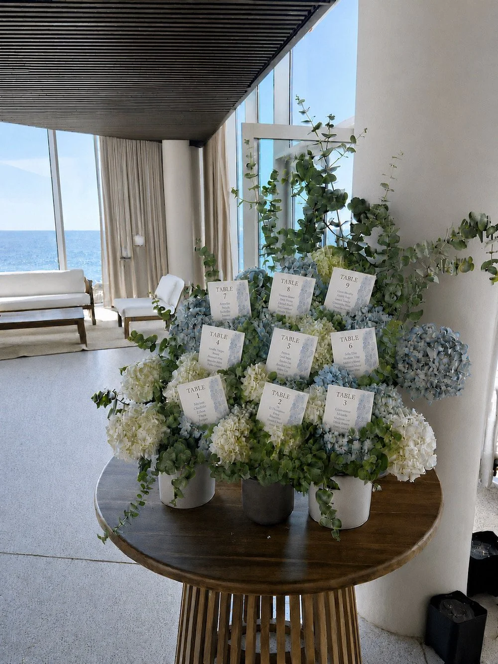 Seating chart with flower arrangements, placed on a round wooden table near large windows showing the ocean, in a bright indoor space.