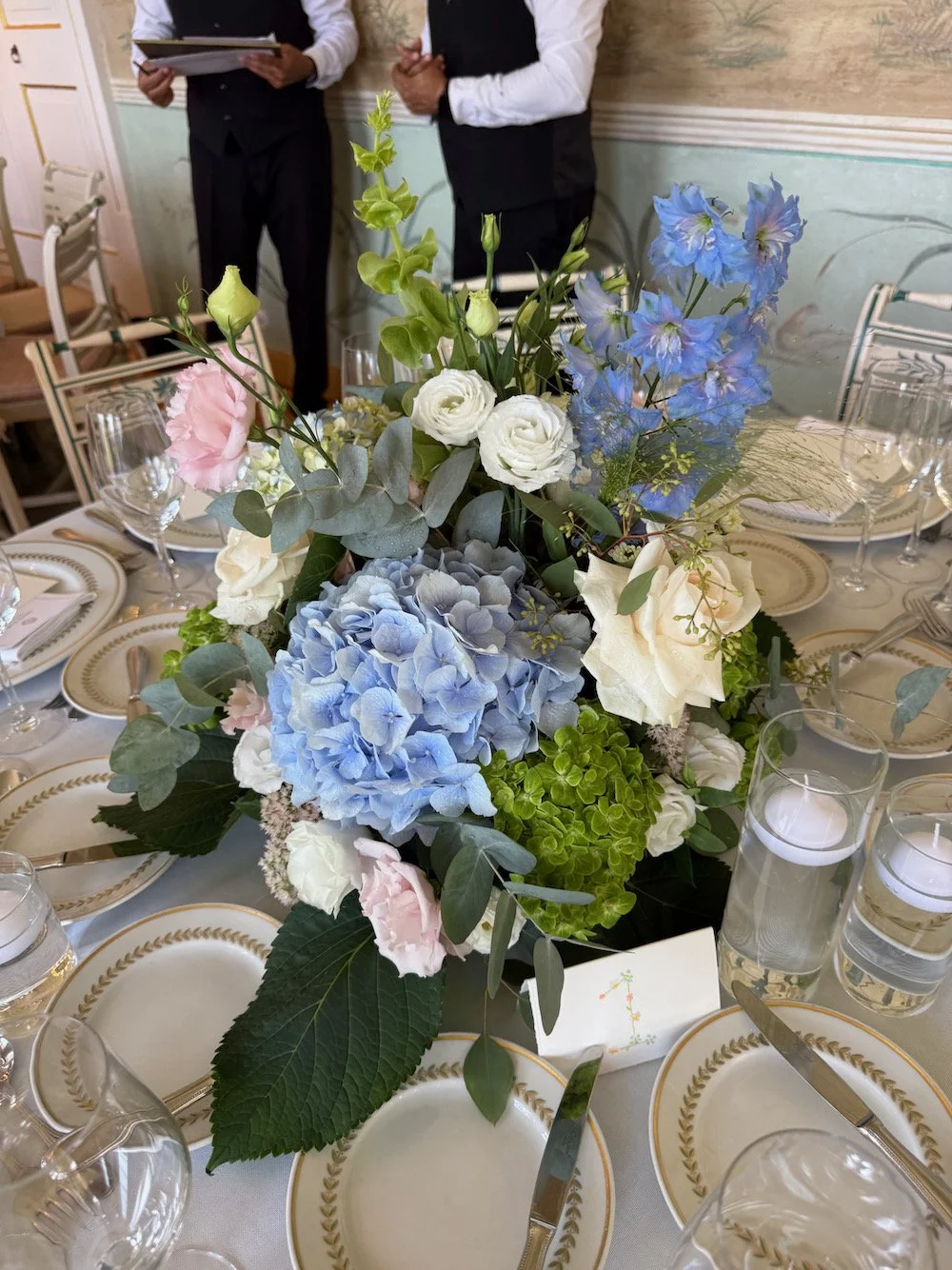 Elegant table setting with a floral centerpiece of blue hydrangeas, white roses, pink lisianthus, and greenery, surrounded by gold-edged plates, clear glasses, and silverware, with two men standing in the background.
