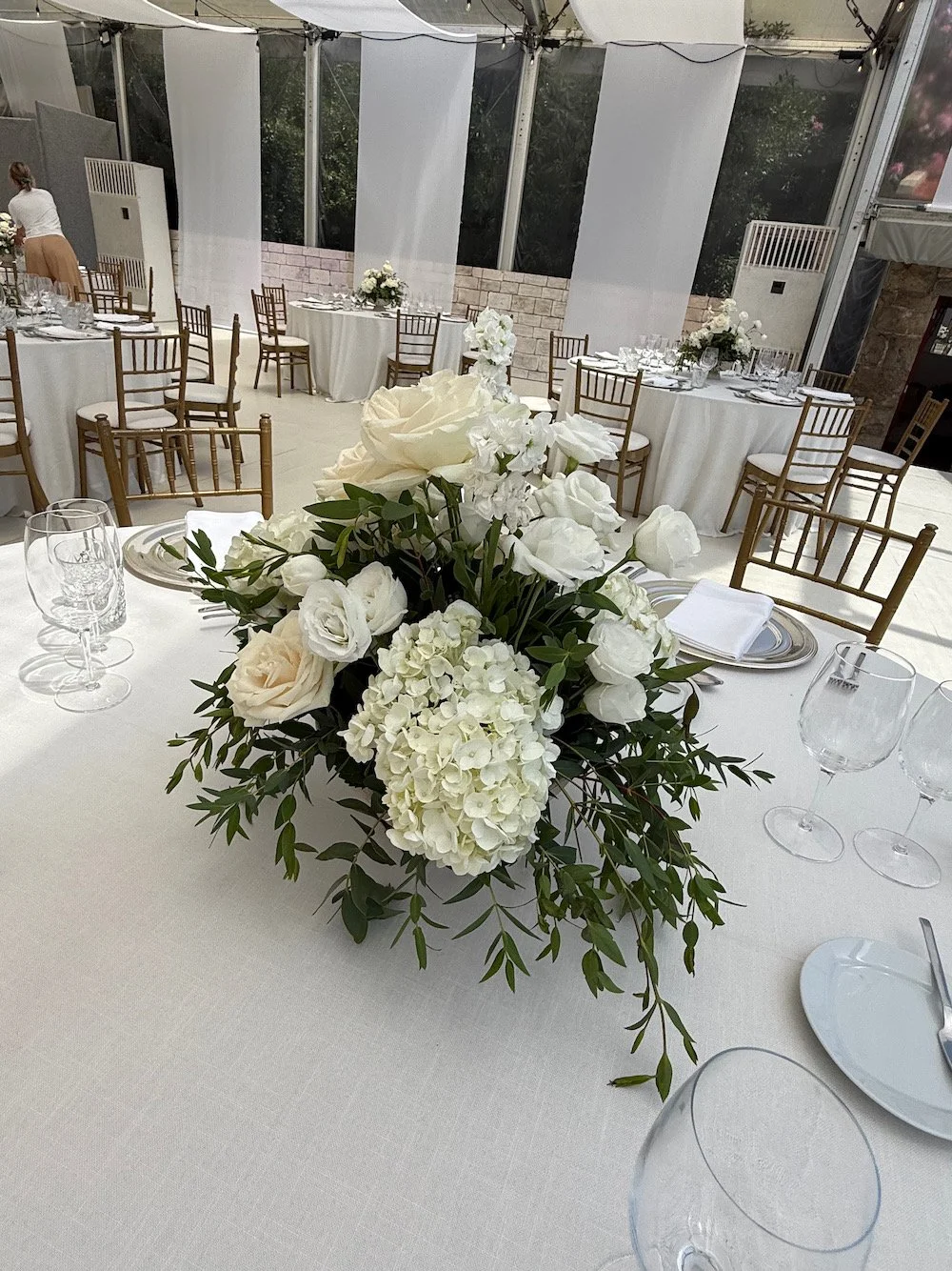 A banquet table decorated with a white tablecloth and a floral centerpiece of white roses, hydrangeas, and greenery, set for an elegant event with plates, silverware, and wine glasses arranged around it.