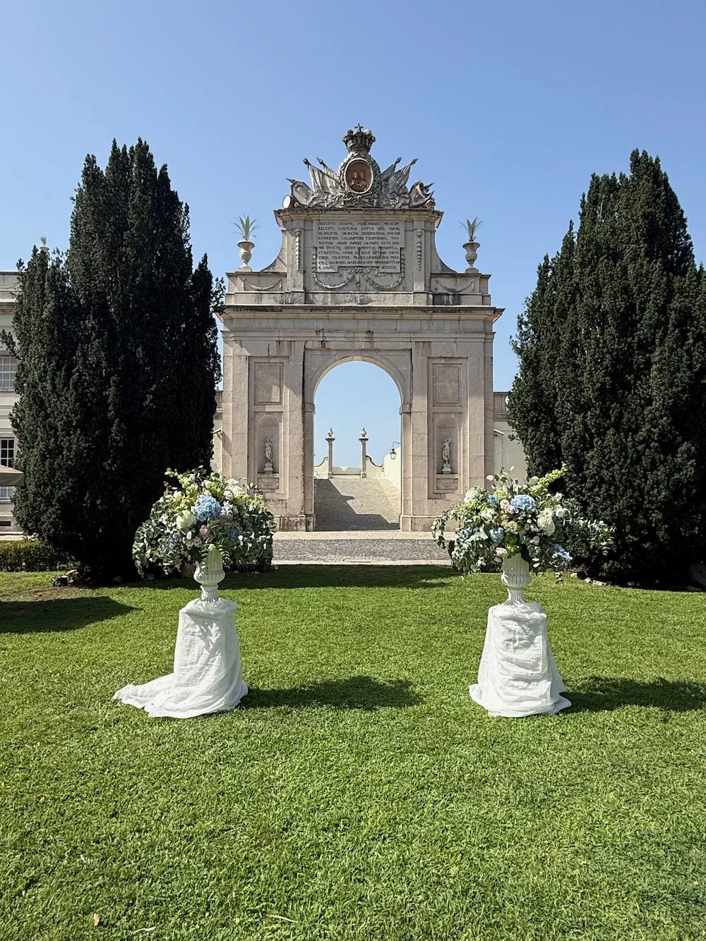 A decorative archway with a Latin inscription and a crest at the top, flanked by two tall trees, with two floral arrangements in white vases on the grass in front, suggests a wedding or special event setting.