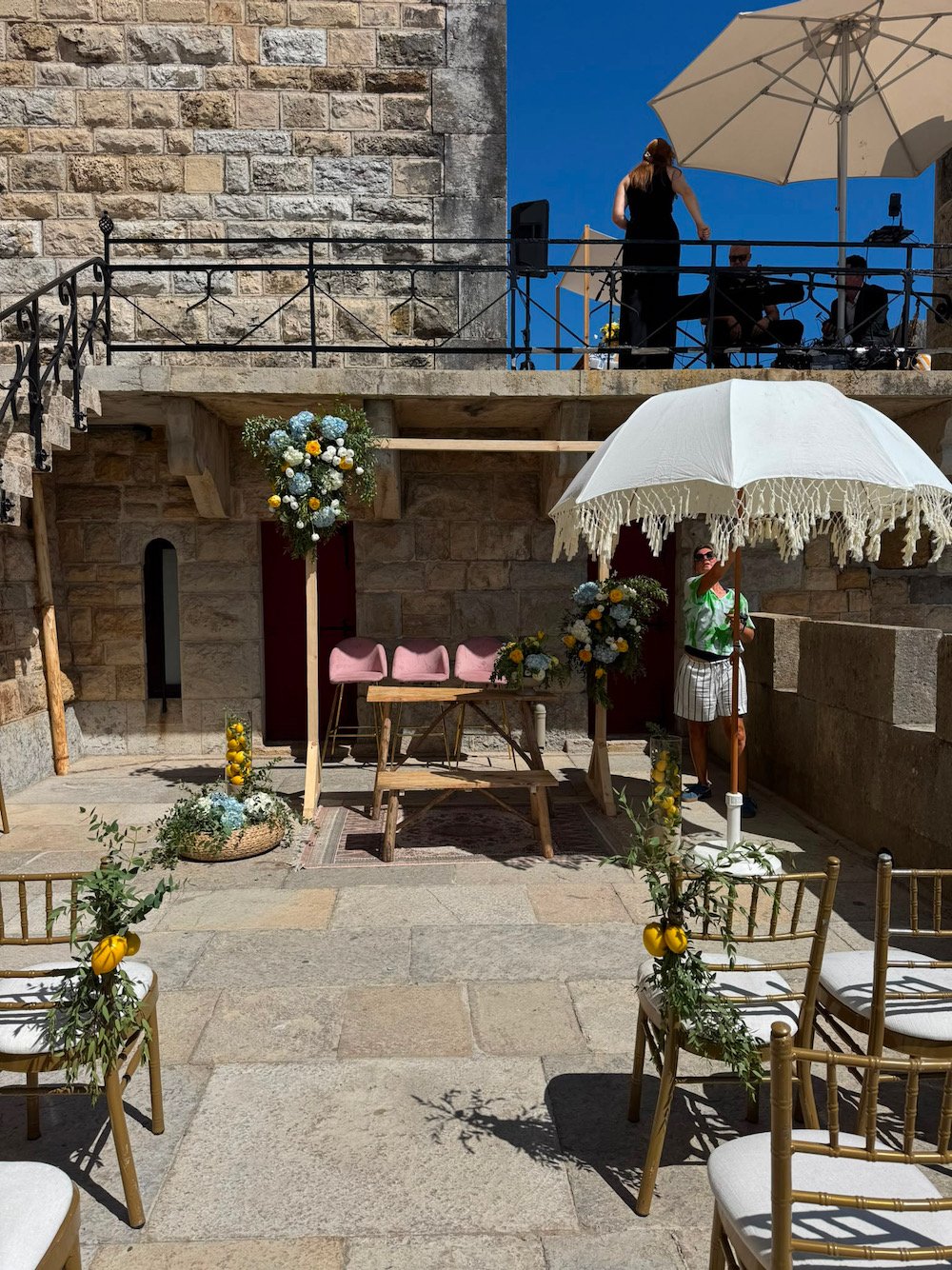 A wedding ceremony setup outdoors with chairs decorated with greenery and lemons, a small table with flowers, a large white umbrella, and a platform with performers and musicians under a large umbrella against a stone wall.