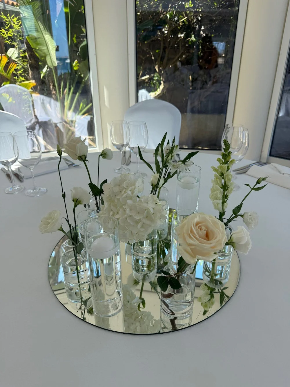 Elegant round table with white floral centerpiece in glass vases on a mirror, surrounded by wine glasses and white chairs, next to large windows with plants outside.
