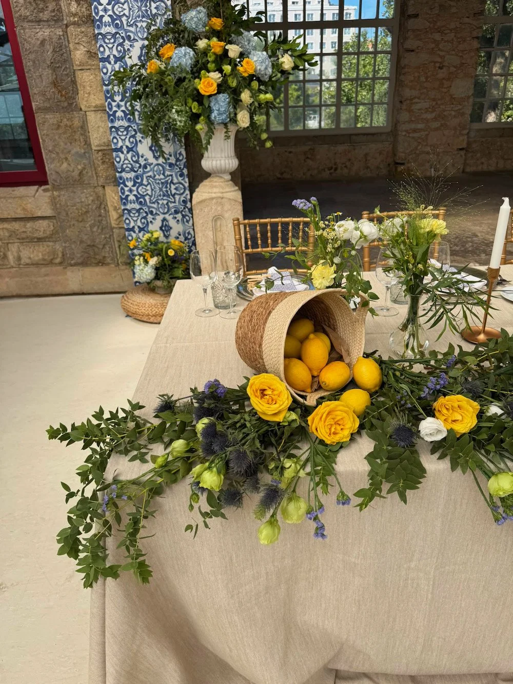 A decorated table with a beige tablecloth, a floral centerpiece with yellow roses, white and purple flowers, and greenery, a straw container with lemons, and tall white candles. In the background, there is a large flower arrangement in a white urn an