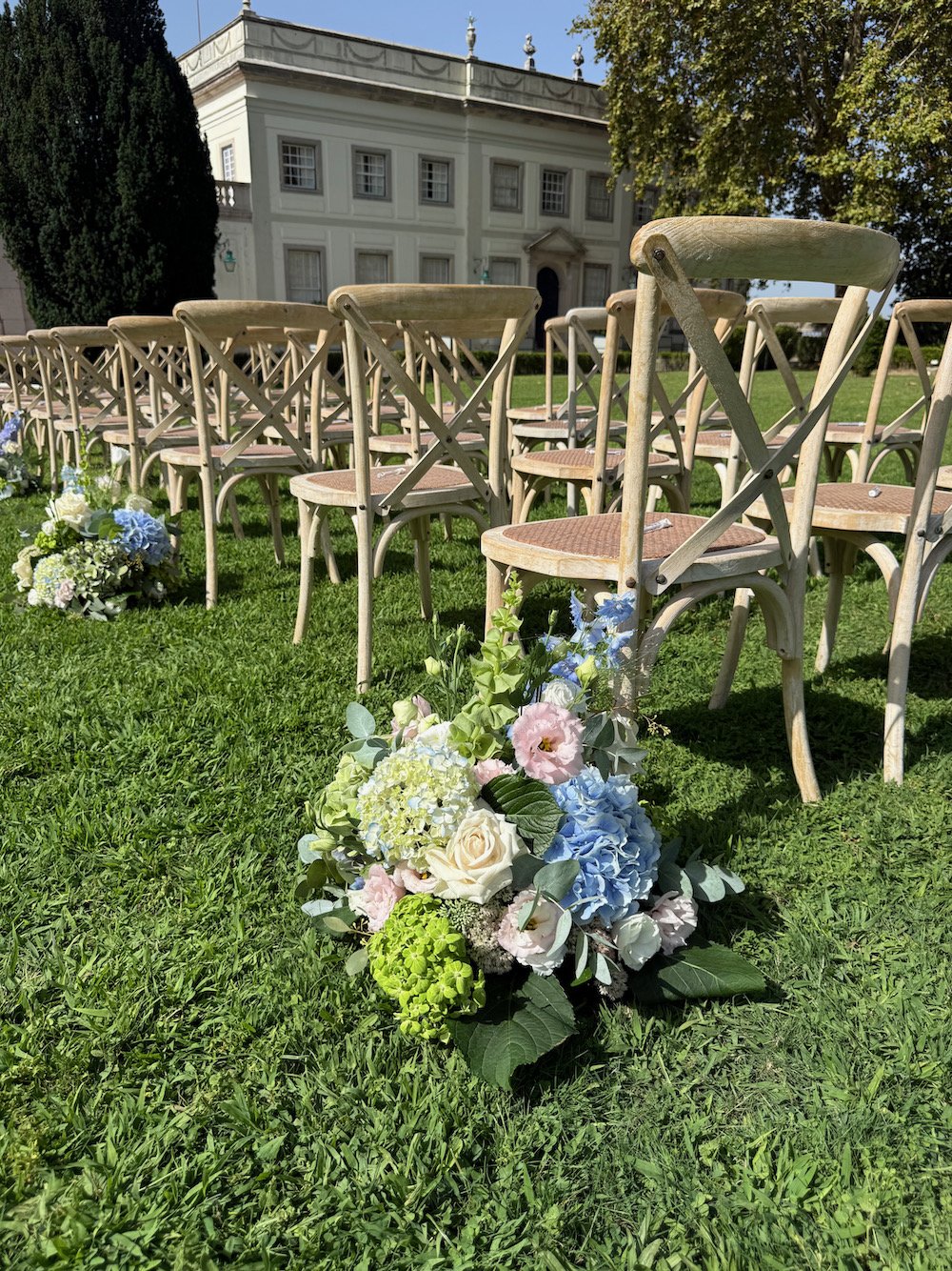Rows of wooden chairs arranged outdoors with floral arrangements on the grass, in front of a white building, suggesting a wedding or outdoor event.