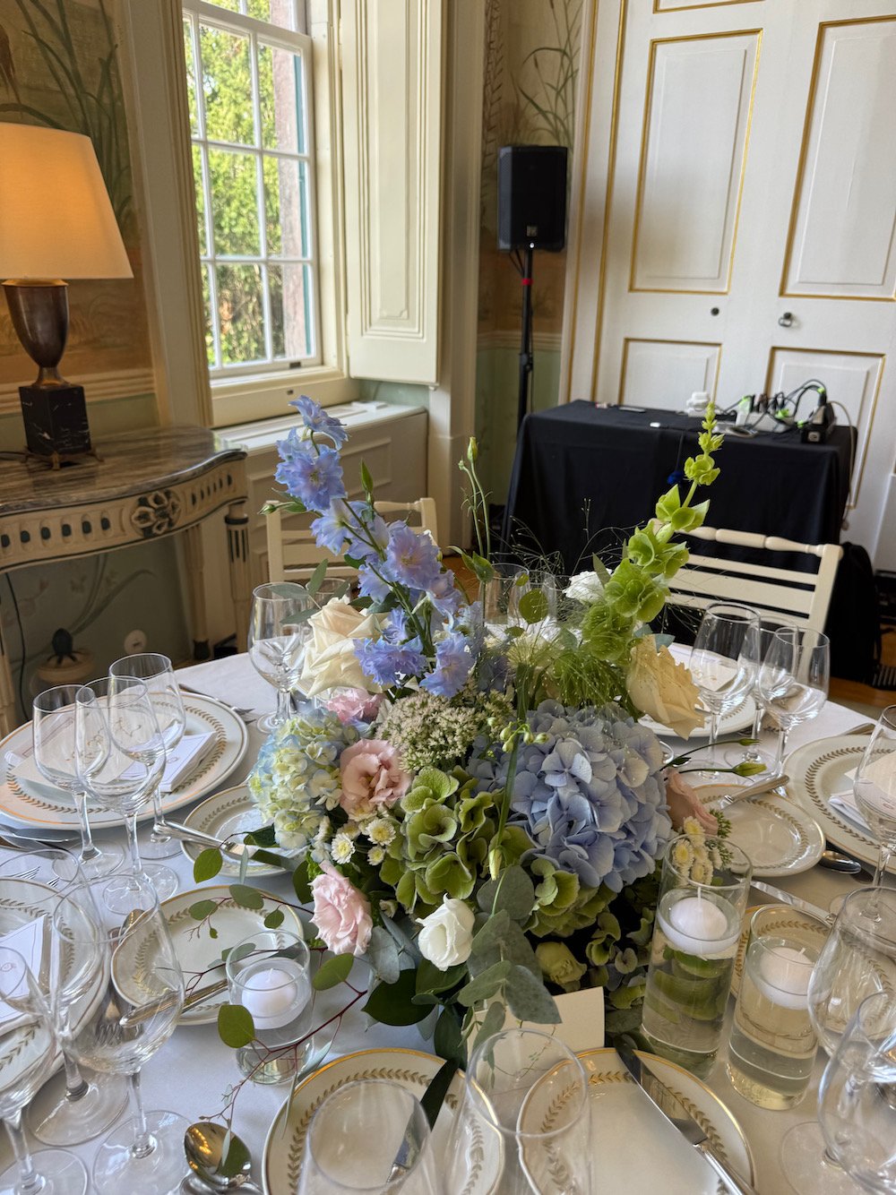 A formal dining table decorated with a large floral centerpiece featuring pastel blue, pink, white, and green flowers. The table is set with white and gold-rimmed plates, multiple wine glasses, and silverware. Candles are placed around the centerpiec