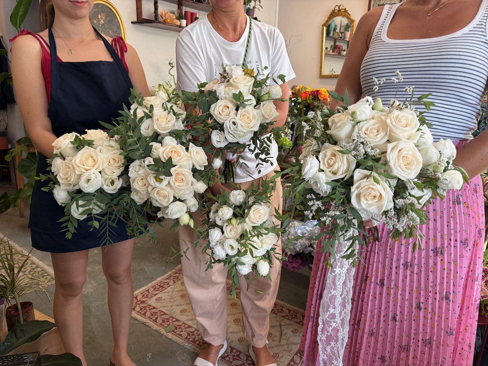 Three women holding large bouquets of white roses and greenery inside a floral shop.