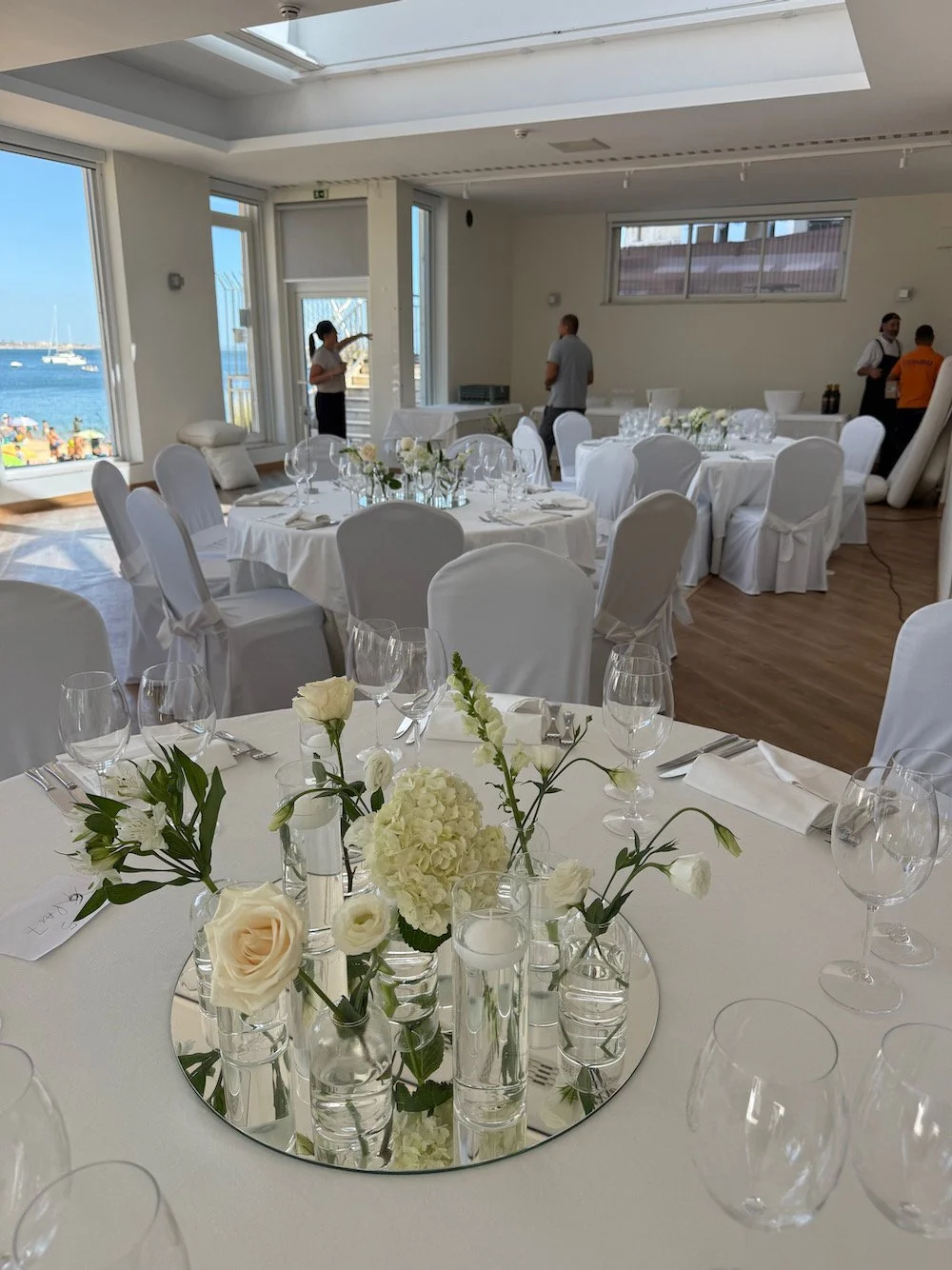 Elegant banquet room set for a special event with white tablecloths, floral centerpieces, and ocean view through large windows.