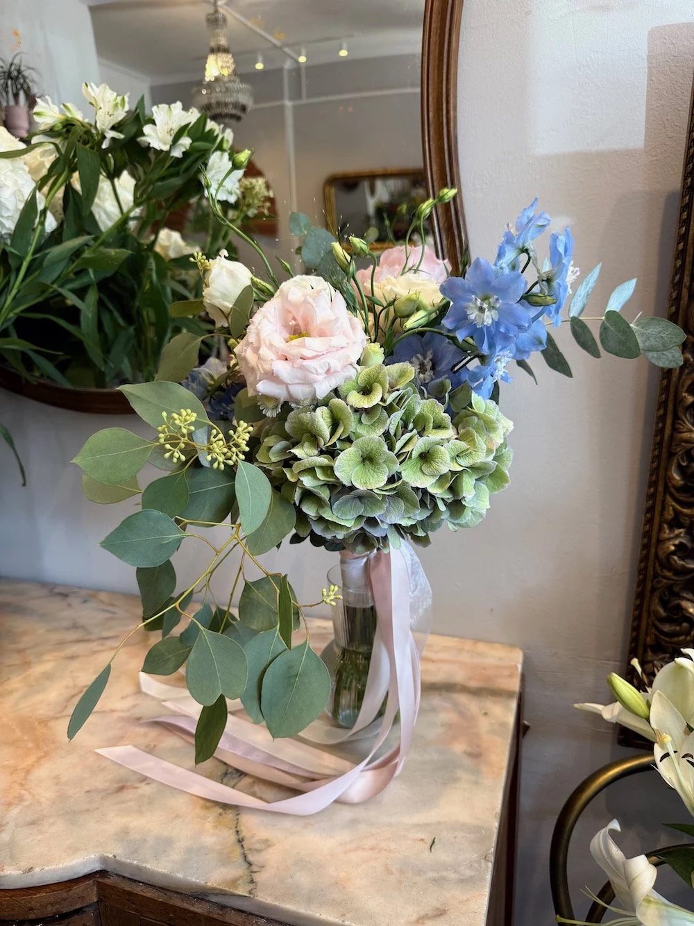 A bouquet of flowers in a glass vase with pink and blue flowers, green leaves, and a white ribbon, placed on a marble table.