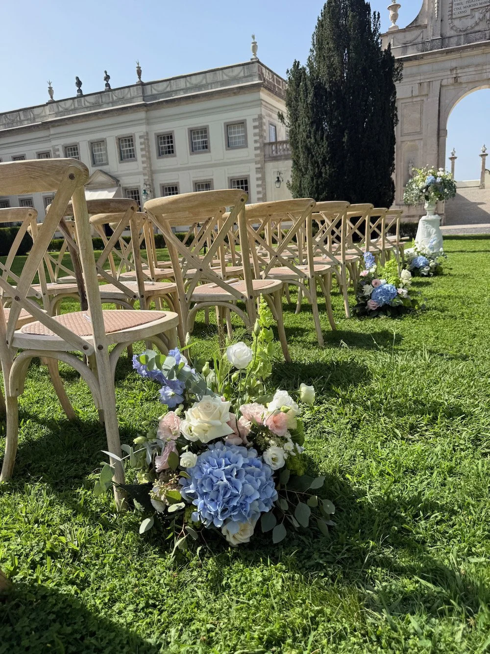 Outdoor wedding setup with wooden chairs arranged on grass, decorated with floral arrangements of white, pink, and blue flowers, in front of a historical building with classical architecture and a blue sky.